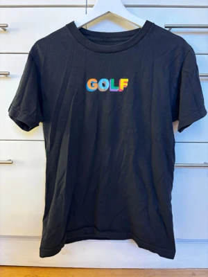 Svart GOLF t-shirt med multifärgat tryck - Svart t-shirt från GOLF med  multifärgat tryck på bröstet. Klassisk rund hals och korta ärmar. Materialet är mjuk bomull och passformen är normal. Storlek S.