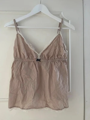  Odd Molly - Supersöt beige-brun linnetopp från Odd Molly i storlek L med tunna axelband och v-ringad hals. Toppen har dekorativ spets längs kanterna och en liten rosett framtill. Broderad Odd Molly-logga med fjädrar på ryggen. Mjukt bomullsmaterial. Säljer den då den tyvärr inte kommer till användning. 