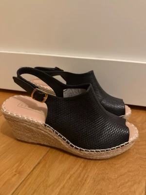 Svarta kilklackade sandaler Style Inc - Svarta sandaler med kilklack från Style Inc i storlek 38. Ovandelen har ett diskret mönster och är gjord i syntetmaterial med öppen tå och justerbart spänne. Sulan är i repimitation som ger en somrig känsla.