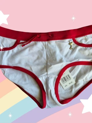 Vit och röd Snobben-trosa från Snoopy XS - Supersöt vit trosa med röda kanter och midja från Snoopy. Framtill finns en liten silvrig Snoopy-berlock och baktill ett rött tryck av Snoopy. Gjord i mjuk bomull och har snörning i midjan för extra detalj. Oanvända från ett varuparti från 2000-talet med lapparna kvar. Bra bundlerabatt om du köper flera varor från mig.