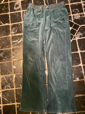 Mörkgröna mjukisbyxor i sammet - Säljer ett par mörkgröna mjukisbyxor från Juicy Couture i mjuk sammet. Byxorna har bred resårmidja med snörning, raka ben och fickor både fram och bak. På ena fickan finns en broderad JC-logga. Perfekta för chill dagar.