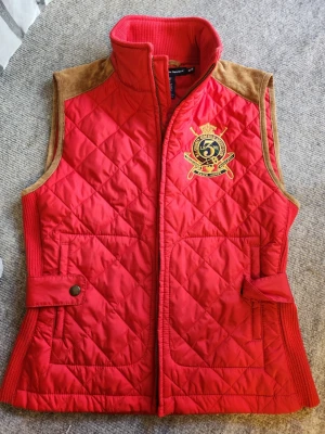 Röd quiltad dunväst från Ralph Lauren - Säljer en snygg röd quiltad dunväst från Ralph Lauren Sport med broderad logga på bröstet. Västen har bruna mockadetaljer vid axlarna, ribbade sidor och hög krage. Materialet är polyester och mocka, med dragkedja framtill och knappar vid midjan. Sparsamt använd