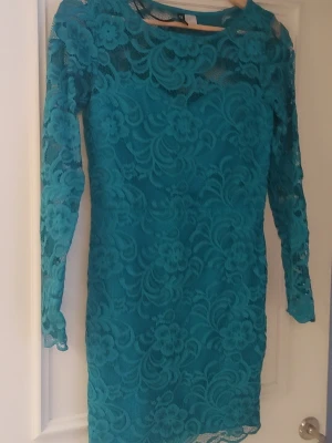 Turkos spetsklänning från H&M - Säljer en turkos kort spetsklänning från H&M med lång ärm och blommigt mönster. Klänningen har en figurnära passform och är helfodrad med transparenta spetsdetaljer på ärmar och över axlarna. Perfekt för dig som vill sticka ut med en färgstark look.