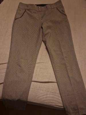 Rutig kostymbyxa från SEZDI, stl 38 - Snygga kostymbyxor från SEZDI i storlek 38. Byxorna har ett rutigt mönster i beige och brunt, två knappar framtill och klassiska fickor. Perfekt för dig som gillar en stilren och trendig look. Mycket bra skick, nästan oanvänd, saknar prislapp. Priset kan diskuteras🫶