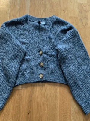 Grå  kofta från H&M  - Mysig croppad kofta från Divided i grått, med grovstickad struktur och tre stora knappar framtill. Koftan har lång ärm och djup v-ringning, perfekt att slänga över en topp för en chill look.