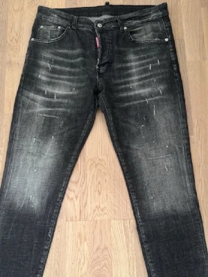 Svarta Dsquared2 jeans med slitningar - Säljer ett par svarta jeans från Dsquared2 med coola slitningar och tvättade detaljer. Modellen har fem fickor, knappgylf och en svart patch med DSQ2 1995 bak. Jeansen har en smal passform och är tillverkade i jeansmaterial med stretch.
