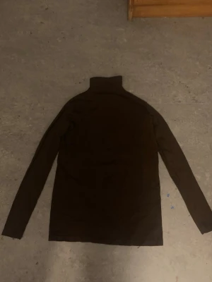 Brun polotröja med lång ärm - Mörkbrun polotröja med lång ärm och hög krage. Tröjan har en enkel och stilren design, perfekt för lager på lager under kyligare dagar. Tillverkad i mjukt material som känns skönt mot huden.