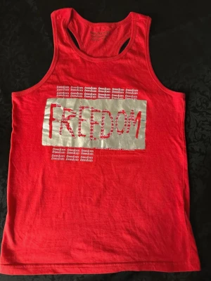 Röd linnetopp från GeeJay med tryck - Cool röd linnetopp från GeeJay med trycket 'FREEDOM' i silver och upprepade små vita 'freedom'-texter. Modellen är ärmlös med brottarrygg och tillverkad i 100% bomull. Perfekt för dig som gillar statement-plagg och sportig stil.