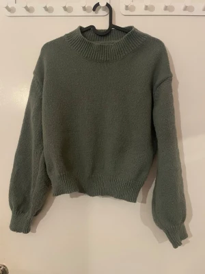 Grön stickad tröja med flätade ärmar - Mysig grön stickad tröja med bred ribbad krage och långa ärmar. Ärmarna har snygga flätade detaljer som ger extra stil. Tröjan har en loose passform och ribbade muddar vid midja och ärmslut. Perfekt för kyliga dagar och enkel att matcha med jeans.