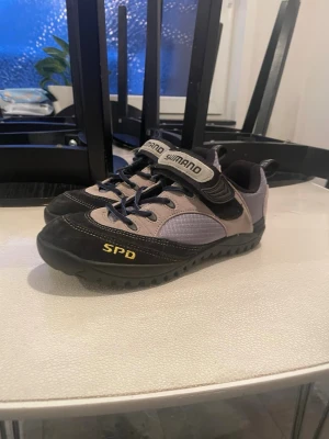 Shimano SPD cykelskor strl 40 - Shimano SPD cykelskor i svart och grått med gula detaljer. Skorna har snörning och kardborreband, robust gummisula med SPD-klossar för cykling. Ovandelen är i mocka och syntetmaterial, perfekt för dig som vill ha grymt grepp och komfort på cykelturen.