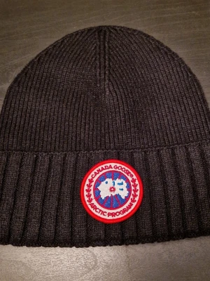 Svart ribbad mössa från Canada Goose - Svart ribbstickad mössa från Canada Goose med klassisk rund logotyp framtill. Mössan är tillverkad i en blandning av ull och polyester, vilket gör den både varm och mjuk. Perfekt för kalla dagar och har en stilren look som funkar till de flesta jackor.