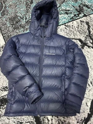 Mörkblå Peak Performance dunjacka - Säljer en mörkblå dunjacka från Peak Performance med huva och dragkedja framtill. Jackan har quiltad design och snörning vid huvan. Perfekt för kalla daga och har en riktigt schysst puffer-look som är både snygg och praktisk.