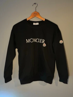 Svart sweatshirt från Moncler - Säljer en svart sweatshirt från Moncler med klassisk logga på bröstet och ärmen. Tröjan har rund halsringning och långa ärmar. Tillverkad i mjuk bomull för skön känsla och stilren look. Perfekt för dig som gillar exklusiva streetwear-plagg.