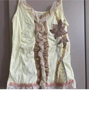 Gul linnetopp med spets från Saint Tropez - Säljer en ljusgul linnetopp från Saint Tropez i storlek S. Toppen har spetsdetaljer vid halsringning och nederkant, volang framtill och en broderad blomma i brunt och beige. Materialet är tunt och luftigt, perfekt för varma dagar.