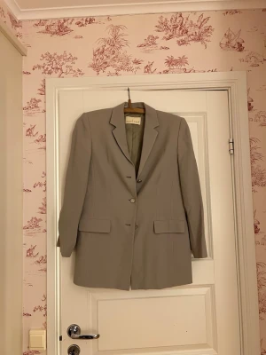 Grå/grön  kavaj från Alexander - Snygg beige kavaj från Akazia House med klassisk krage och tre knappar framtill. Kavajen har två fickor med lock och är långärmad.