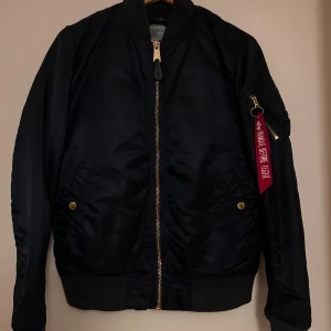 Mörkblå bomberjacka Alpha Industries L - Säljer en klassisk mörkblå bomberjacka från Alpha Industries med guld dragkedjor och detaljer. Jackan har ribbade muddar, fickor med tryckknappar och ikonisk röd tag på ärmen. Tillverkad i slitstark polyester och har en snygg, tidlös look.