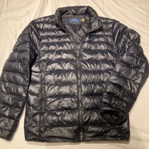 Svart pufferjacka från Polo Ralph Lauren - Snygg glansig svart pufferjacka från Polo Ralph Lauren med klassisk broderad logga på bröstet. Jackan har dragkedja framtill, två sidofickor med dragkedja och quiltad design. Perfekt för kyliga dagar och riktigt skön att bära. Skriv om ni har frågor.
