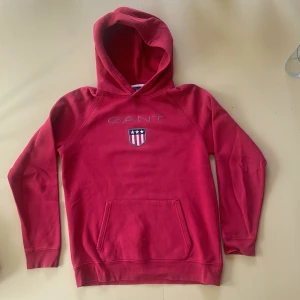 Röd hoodie från GANT med logga - Röd hoodie från GANT med broderad logga och sköld på bröstet. Klassisk känguruficka framtill och huva. Mjuk bomullskänsla och långa ärmar, perfekt för chill dagar. Snygg och enkel design som funkar till det mesta.
