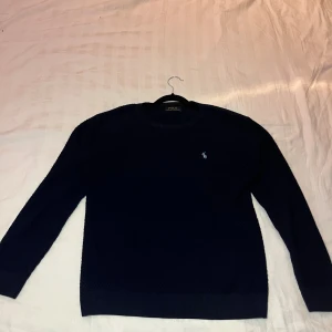 Mörkblå stickad tröja Polo Ralph Lauren - Säljer en mörkblå stickad tröja från Polo Ralph Lauren. Klassisk modell med rund halsringning, diskret broderad logga på bröstet och ribbade muddar vid ärmslut och nederkant. Perfekt för dig som gillar stilrena och tidlösa plagg. dock så är den lite knottrigt