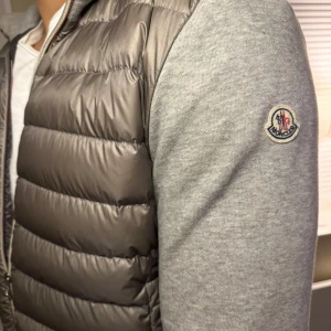 Grå dunväst från Moncler - Snygg grå dunväst från Moncler med quiltad front i glansigt material och mjuka grå ärmar. Västen har dragkedja framtill och klassisk Moncler-logga på ärmen. Perfekt för lager-på-lager och streetstyle.