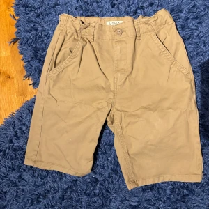 Beiga chino shorts från Lindex - Snygga beiga shorts från Lindex i storlek 164. Klassisk modell med fickor fram och bak, stängs med knapp och dragkedja. Perfekta för varma dagar och har en avslappnad passform. Tillverkade i mjuk bomull för skön känsla.
