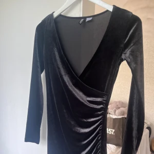 Svart sammetsklänning från H&M Divided - Supersnygg svart sammetsklänning från H&M Divided i omlottmodell med v-ringning och långa ärmar. Klänningen är kort och har snygg rynkdetalj längs sidan som ger en cool och trendig look. Perfekt för dig som vill sticka ut lite extra.