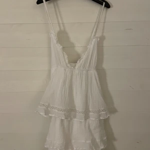 Vit somrig klänning från Gina Tricot - Superfin vit kort klänning från Gina Tricot med tunna axelband som korsas i ryggen. Klänningen har volangdetaljer, spetsinslag och en luftig, lätt struktur. Perfekt för varma dagar och ger en romantisk vibe.