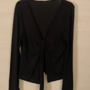 Svart omlottopp med knyt framtill - Snygg svart omlottopp med djup v-ringning och långa ärmar. Toppen har ett knyt framtill som ger en cool och trendig look. Perfekt att styla med jeans eller kjol för en stilren outfit.