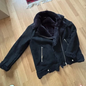 Svart teddyjacka med dragkedja - Snygg svart teddyjacka med mjukt foder och stora slag. Jackan har silverfärgade dragkedjor och detaljer vid ärmslut och nederkant. Perfekt för kalla dagar och ger en cool look.