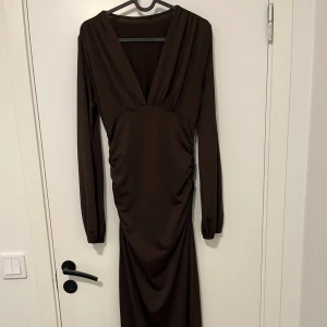 Brun långklänning med v-ringning - Säljer en elegant brun långklänning med djup v-ringning och långa ärmar. Klänningen har snygga rynkade detaljer vid midjan som ger en smickrande siluett. Perfekt för dig som gillar stilrena och sofistikerade looks.