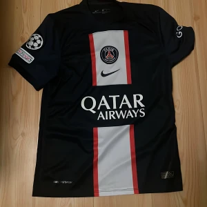 PSG Messi #30 Nike fotbollströja S - Paris Saint-Germain matchtröja med Messi #30 på ryggen. Tröjan är svart med breda vita och röda vertikala ränder, PSG-emblem och Qatar Airways-tryck på bröstet. Champions League-märke på ärmen. Tillverkad i Nike VaporKnit-material för bästa komfort.
