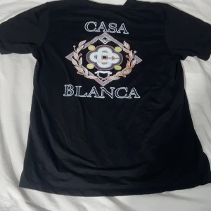 Svart Casa Blanca t-shirt med tryck - Svart t-shirt från Casa Blanca med stort färgglatt emblem och text på ryggen samt mindre tryck på bröstet. Klassisk passform och rund hals. Tillverkad i mjuk bomull, perfekt för en clean och stilren look.