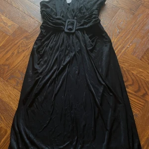 Svart långklänning från Mango Suit - Elegant svart långklänning från Mango Suit i glansig polyester. Klänningen har markerad midja med bredt bälte och snygga rynkade detaljer vid axlar och sidor. Djup v-ringning framtill och dragkedja i sidan för enkel påklädning. Perfekt för dig som vill sticka ut.