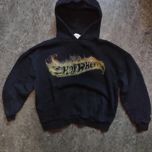  Pull&Bear Hot Wheels  - Svart hoodie från Pull&Bear i samarbete med Hot Wheels. Stor logo med eld-detaljer i gult och orange framtill. Klassisk huva och känguruficka. Perfekt för dig som gillar streetwear och coola prints.