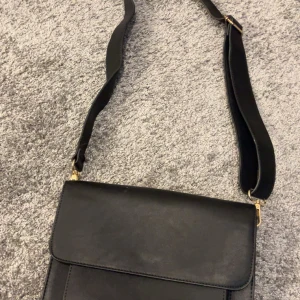 Svart axelväska i skinn - Stilren svart axelväska med bred justerbar rem och guldfärgade detaljer. Väskan har en enkel, fyrkantig form och lockstängning. Perfekt för dig som vill ha en klassisk och snygg accessoar till vardagslooken.