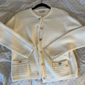 Vit kofta från Holly & Whyte XS - Snygg vit kofta från Holly & Whyte med guldknappar och två fickor framtill. Koftan har ett diskret strukturerat mönster och är långärmad med rund hals. Perfekt att slänga över en topp för en clean och fräsch look. Koftan är använd mycket få gånger. Tveka inte att höra av dig!