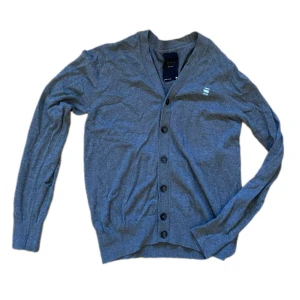 Grå kofta från G-Star RAW - Grå kofta från G-Star RAW med knappar framtill och diskret logga på bröstet. Klassisk v-ringning och långärmad modell i mjuk bomull. Perfekt för lager-på-lager och enkel att matcha med andra plagg.