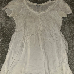 Vit blus med knyt från Gina Tricot - Söt vit blus från Gina Tricot i storlek 38. Blusen har korta ärmar, rynkade detaljer och knyt framtill vid halsen. Den är tillverkad i ett lätt och luftigt tyg, perfekt för varmare dagar. Rundad halsringning med volangkant ger en romantisk känsla.