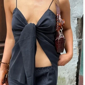 Svart omlottopp från Djerf Avenue - Supersnygg svart omlottopp från Djerf Avenue med tunna axelband och stor knytning framtill. Toppen har en croppad passform och är tillverkad i ett mjukt, slätt material som ger en lyxig känsla. Perfekt för att skapa en trendig och stilren look.