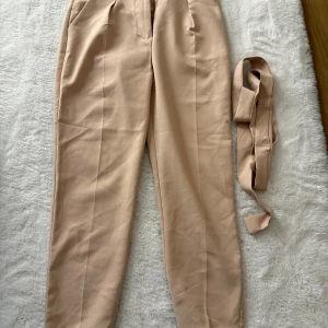 Beiga kostymbyxor med bälte - Snygga beiga kostymbyxor med tillhörande bälte. Byxorna har hög midja, raka ben och pressveck framtill för en clean look. Perfekta för dig som gillar stilrena och klassiska plagg. Materialet känns mjukt och följsamt.