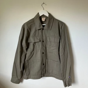 Olivgrön overshirt från Champion - Olivgrön overshirt från Champion i 100% bomull. Jackan har raka linjer, klassisk krage, fyra stora fickor framtill och knäppning med svarta knappar. Liten broderad Champion-logga på bröstet och ärmslutet. Perfekt lager-på-lager-plagg för en clean streetstil. 3U13
