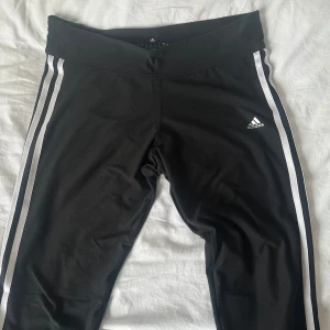 Svarta Adidas träningsbyxor med vita ränder - Svarta leggings från Adidas med klassiska vita ränder längs sidorna och Adidas-logga på låret. Modellen har bred midja och är gjord i stretchigt material som andas, perfekt för träning eller chill. Snygg och sportig look