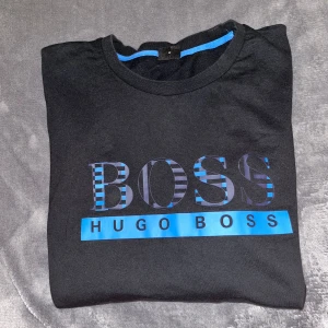 Svart långärmad tröja Hugo Boss - Svart långärmad tröja från Hugo Boss med blått och randigt BOSS-tryck framtill. Tröjan har rund halsringning och blå detalj på insidan av kragen. Perfekt för dig som gillar stilrena och snygga loggoplagg. Endast använd fåtal gånger. Riktigt bekväm och fint material! 