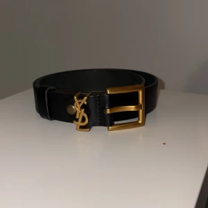 Svart YSL bälte i skinn med gulddetaljer - Snyggt svart bälte från YSL i äkta skinn med klassiskt YSL-logo i guld och matchande guldfärgat spänne. Perfekt accessoar för att lyfta vilken outfit som helst med en lyxig touch.