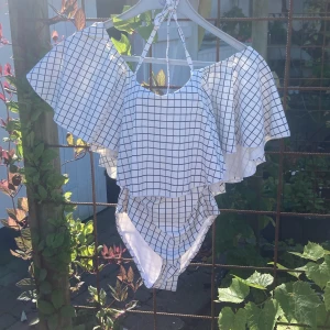 Vit rutig baddräkt med volang - Säljer en superfin baddräkt i vitt med svart rutigt mönster. Bikinin har breda volangärmar och axelband som knyts i nacken. Toppen har en snygg omlottkänsla och ger en unik look på stranden. Perfekt för dig som vill sticka ut i sommar!