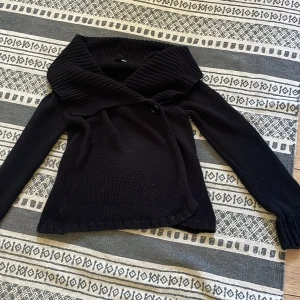 Svart stickad kofta från H&M - Svart stickad kofta från H&M med stor, ribbad krage och omlottknäppning med knapp. Långärmad och i mjukt material som passar perfekt när det är kyligt. Snygg och enkel att matcha med olika outfits.