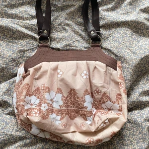 Blommig axelväska i beige och brunt - Söt axelväska med blommigt mönster i vitt och brunt på beige botten. Väskan har bruna handtag i skinnimitation och rundad form. Perfekt för dig som vill ha en unik och charmig accessoar med plats för det viktigaste.