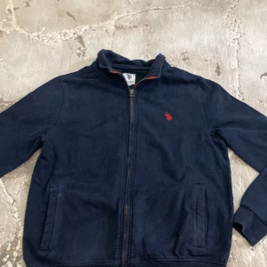 Marinblå ziptröja från U.S. Polo Assn. - Marinblå långärmad ziptröja från U.S. Polo Assn. med röd broderad logga på bröstet. Tröjan har ståkrage, dragkedja framtill och två fickor. Perfekt för lager-på-lager och chill stil.