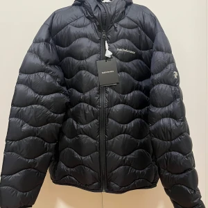 Svart dunjacka Peak Performance XL - Svart dunjacka från Peak Performance i storlek XL. Jackan har vågformat quiltat mönster, huva och dragkedja framtill. Perfekt för kalla dagar och riktigt snygg streetstil. Broderad logga på bröstet och lätt glansigt material. Ordinarepris är 2999.