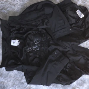 Svart Adidas vindjacka 808 Nachtklub - Svart vindjacka från Adidas med stort broderat 808 Nachtklub Berlin-tryck på ryggen. Jackan har klassisk krage, tryckknappar och är tillverkad i lätt polyester. Perfekt för dig som gillar streetwear och vill sticka ut med en unik detalj.